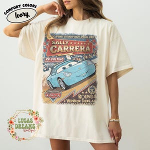 May include: Ivory-colored t-shirt featuring a graphic of Sally Carrera from the Disney Pixar Cars movie. The design includes the text "SALLY CARRERA" and "ROUND 4 WINNER TAKES ALL". The shirt is from the Comfort Colors brand.