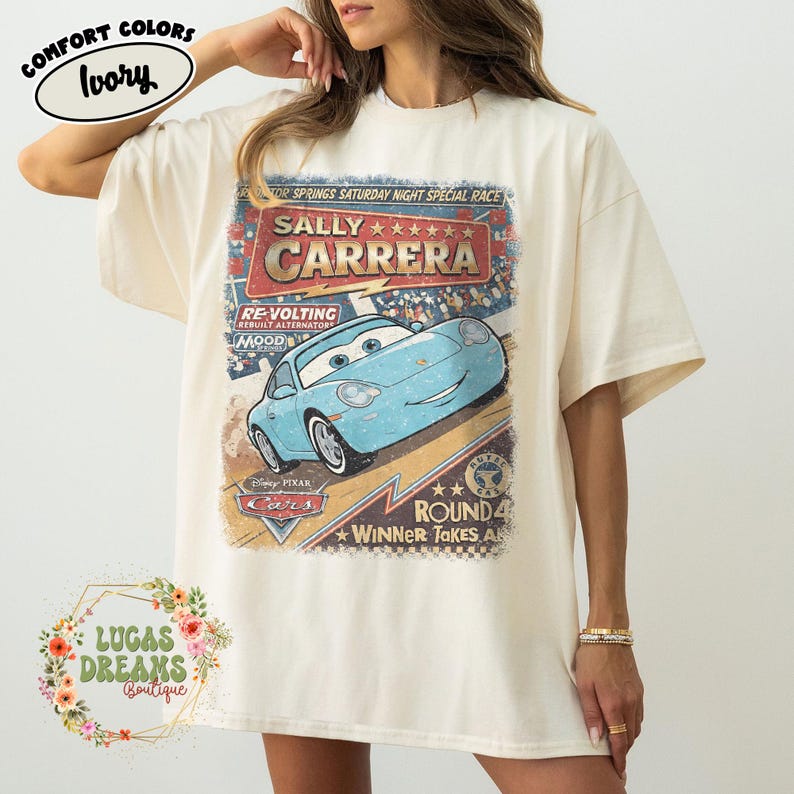 May include: Ivory-colored t-shirt featuring a graphic of Sally Carrera from the Disney Pixar Cars movie. The design includes the text "SALLY CARRERA" and "ROUND 4 WINNER TAKES ALL". The shirt is from the Comfort Colors brand.