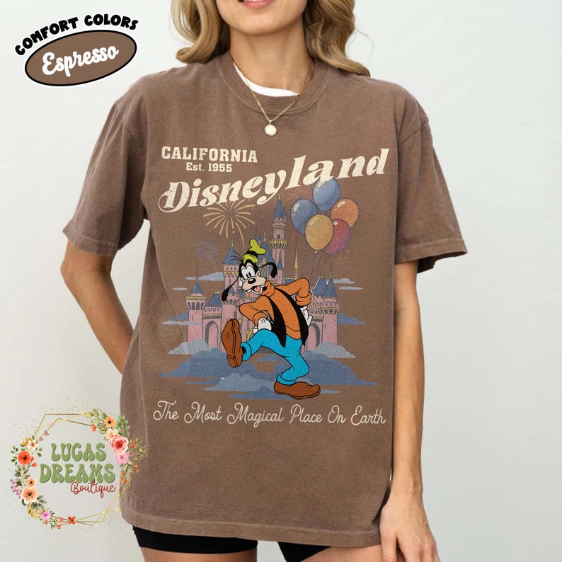 May include: Espresso-colored t-shirt featuring a Disneyland graphic. The design includes Goofy, balloons, a castle, and the text "California Est. 1955 Disneyland" and "The Most Magical Place On Earth."