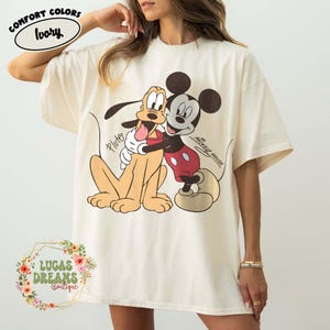 Puede incluir: Camiseta Comfort Colors color marfil con un gráfico de Mickey Mouse abrazando a Pluto. Los textos "Mickey Mouse" y "Pluto" son visibles. La camiseta es de Lucas Dreams Boutique.