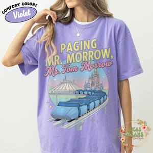 May include: Violet Comfort Colors t-shirt featuring the text "Paging Mr. Morrow, Mr. Tom Morrow" above a graphic of a roller coaster, Space Mountain, and a castle. The shirt has a crew neck and short sleeves.