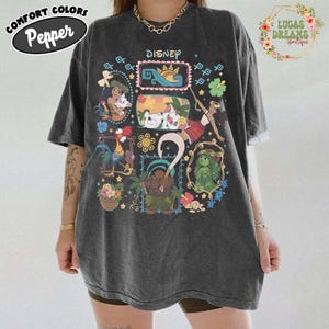 May include: Dark grey oversized t-shirt featuring Disney characters in colourful frames. The shirt has the word "Disney" printed at the top and the words "Comfort Colors Pepper" in a circular logo. The shirt is worn by a person.