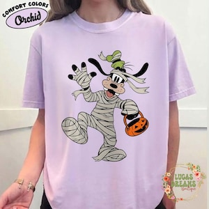 May include: A light purple t-shirt featuring a cartoon character dressed as a mummy, holding a jack-o'-lantern bucket. The shirt has the words "Comfort Colors" and "Orchid" printed on it. The shirt also has the words "LUCAS DREAMS Boutique" printed on it.