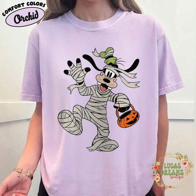 May include: A light purple t-shirt featuring a cartoon character dressed as a mummy, holding a jack-o'-lantern bucket. The shirt has the words "Comfort Colors" and "Orchid" printed on it. The shirt also has the words "LUCAS DREAMS Boutique" printed on it.