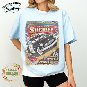May include: Light blue t-shirt featuring a graphic of Sheriff from Disney Pixar's Cars. The design includes the text "RADIATOR SPRINGS SATURDAY NIGHT SPECIAL RACE" and "ROUND 4 WINNER TAKES ALL". The shirt has the Comfort Colors Chambray label.