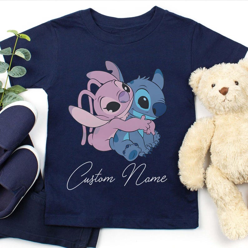 May include: Navy blue t-shirt featuring a graphic of two cartoon characters hugging, one pink and one blue. The text "Custom Name" is written in cursive below the graphic. A teddy bear and a pair of blue shoes are also in the image.
