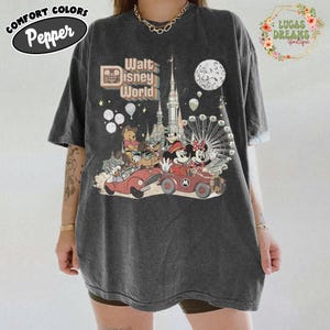 May include: Dark gray t-shirt with a colorful graphic featuring Disney characters in cars, a castle, and a Ferris wheel. The text "Walt Disney World" is displayed above the characters. The shirt also has the text "Comfort Colors Pepper" on the top left.