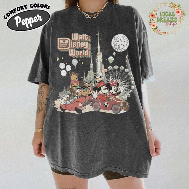 May include: Dark gray t-shirt with a colorful graphic featuring Disney characters in cars, a castle, and a Ferris wheel. The text "Walt Disney World" is displayed above the characters. The shirt also has the text "Comfort Colors Pepper" on the top left.