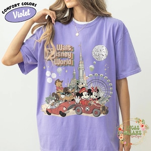 May include: Violet Comfort Colors t-shirt featuring a colorful graphic of Disney characters in cars, with a castle, Ferris wheel, and moon. The text "Walt Disney World" is above the graphic. The shirt has a crew neck.
