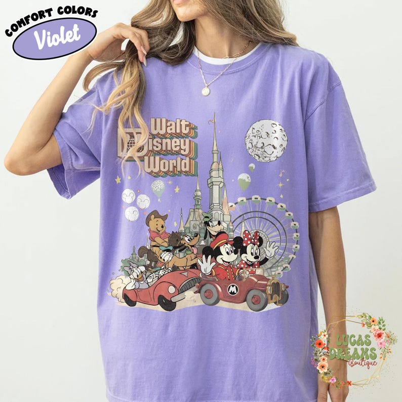 May include: Violet Comfort Colors t-shirt featuring a colorful graphic of Disney characters in cars, with a castle, Ferris wheel, and moon. The text "Walt Disney World" is above the graphic. The shirt has a crew neck.