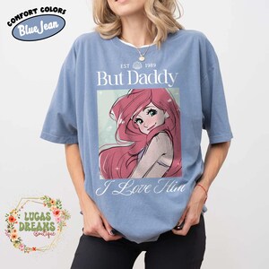 May include: Blue short-sleeved t-shirt with the text "But Daddy I Love Him" and an illustration of a cartoon character with long red hair. The shirt also has the text "EST 1989" and the words "Comfort Colors Blue Jean".