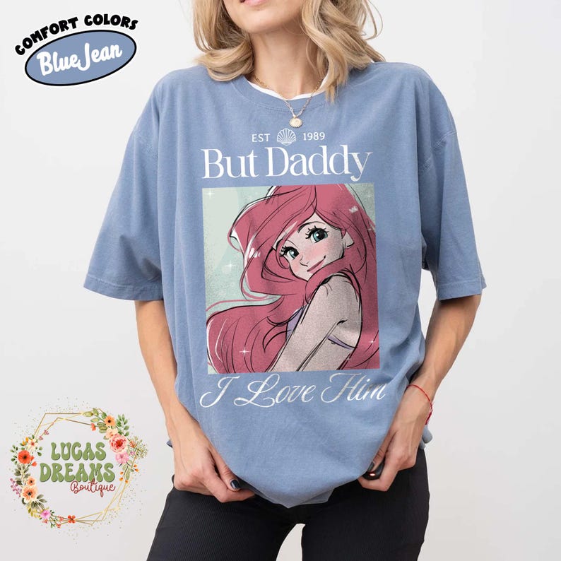 May include: Blue short-sleeved t-shirt with the text "But Daddy I Love Him" and an illustration of a cartoon character with long red hair. The shirt also has the text "EST 1989" and the words "Comfort Colors Blue Jean".
