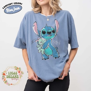May include: A blue short-sleeve t-shirt featuring a graphic illustration of the character Stitch holding a small doll. The design includes the text Stitch in a stylized script. The shirt has a crew neckline and a relaxed, loose-fitting silhouette.
