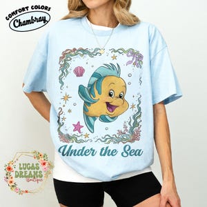 May include: Light blue t-shirt featuring a cartoon illustration of a yellow and blue fish with the text "Under the Sea". The shirt has the words "Comfort Colors" and "Chambray" printed on the top left.
