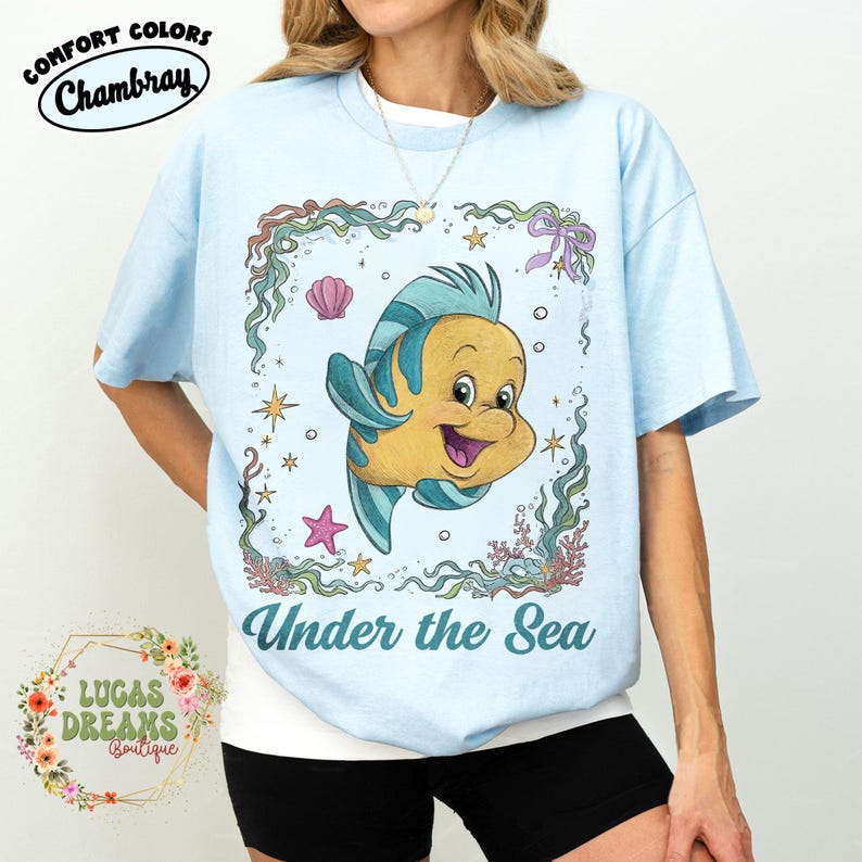 May include: Light blue t-shirt featuring a cartoon illustration of a yellow and blue fish with the text "Under the Sea". The shirt has the words "Comfort Colors" and "Chambray" printed on the top left.