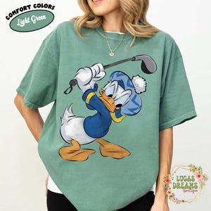 May include: Light green comfort colors t-shirt featuring a cartoon Donald Duck character playing golf. Donald Duck is wearing a blue hat and sweater with yellow accents. The shirt has the text "COMFORT COLORS Light Green".