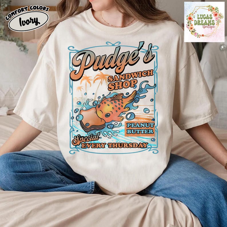 Pu&ograve; includere: T-shirt Comfort Colors color avorio con grafica per "Pudge's Sandwich Shop". Il design include un sandwich di pesce dei cartoni animati, palme e la scritta "Peanut Butter Special Every Thursday."