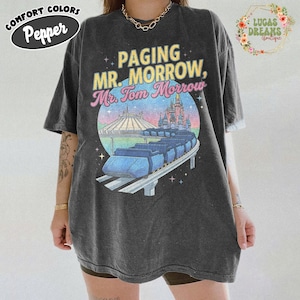 May include: Dark grey t-shirt with a graphic featuring the text "Paging Mr. Morrow, Mr. Tom Morrow" above a colourful illustration of a monorail, Space Mountain, and a castle. The shirt has the words "Comfort Colors" and "Pepper" on it.