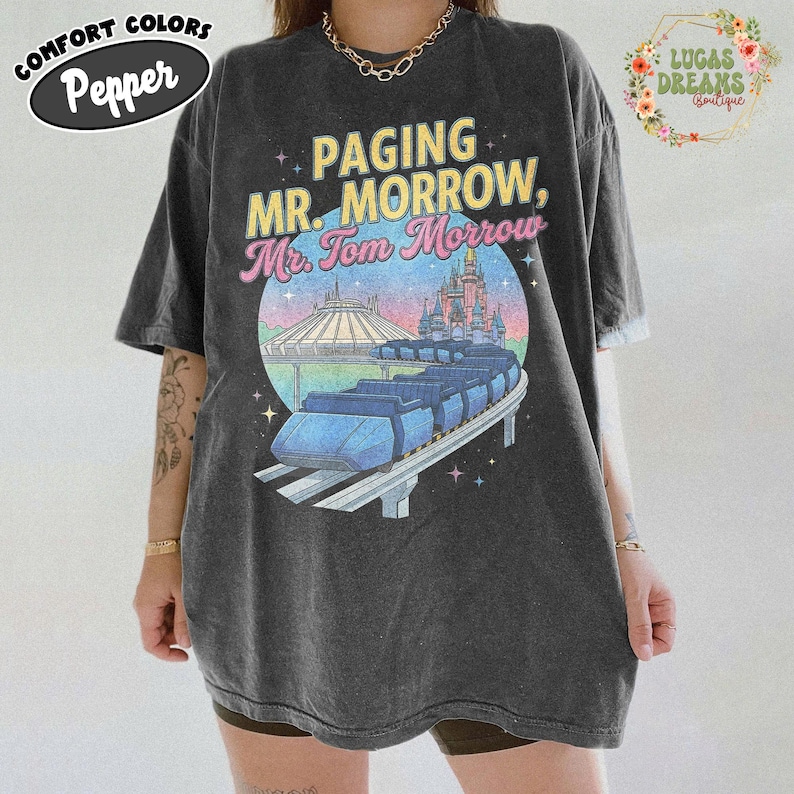May include: Dark grey t-shirt with a graphic featuring the text "Paging Mr. Morrow, Mr. Tom Morrow" above a colourful illustration of a monorail, Space Mountain, and a castle. The shirt has the words "Comfort Colors" and "Pepper" on it.