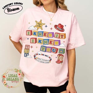 May include: A light pink t-shirt featuring a graphic print of colorful alphabet blocks spelling I KNEW IT I KNEW YOU, a sheriff badge with the text INFINITY AND BEYOND, a red cowboy hat, a beaded bracelet with the text YOU'VE GOT A FRIEND IN ME, and boots.