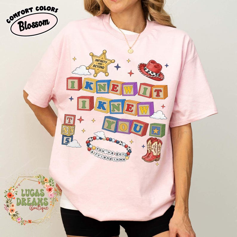 May include: A light pink t-shirt featuring a graphic print of colorful alphabet blocks spelling I KNEW IT I KNEW YOU, a sheriff badge with the text INFINITY AND BEYOND, a red cowboy hat, a beaded bracelet with the text YOU'VE GOT A FRIEND IN ME, and boots.
