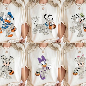 May include: Six cream-colored t-shirts featuring Halloween-themed cartoon characters. Donald Duck, Pluto, and Goofy are wrapped in mummy bandages. Mickey and Minnie Mouse, and Daisy Duck are also in mummy costumes, each holding a jack-o'-lantern bucket.