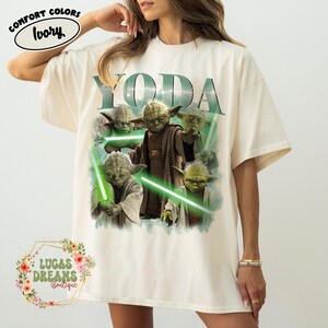 Puede incluir: Una camiseta de color marfil con un estampado gr&aacute;fico del personaje Yoda en varias poses con sables de luz verdes. El texto YODA est&aacute; impreso en letras verdes grandes sobre el personaje. Una peque&ntilde;a etiqueta ovalada indica COMFORT COLORS IVORY.