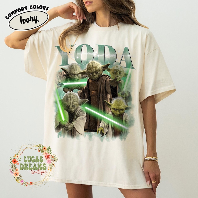 Puede incluir: Una camiseta de color marfil con un estampado gr&aacute;fico del personaje Yoda en varias poses con sables de luz verdes. El texto YODA est&aacute; impreso en letras verdes grandes sobre el personaje. Una peque&ntilde;a etiqueta ovalada indica COMFORT COLORS IVORY.