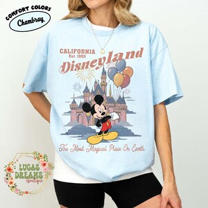 May include: Light blue t-shirt featuring a graphic of Mickey Mouse in front of a castle with balloons. The text reads "CALIFORNIA Disneyland Est. 1955" and "The Most Magical Place On Earth". The shirt has the "Comfort Colors Chambray" logo.