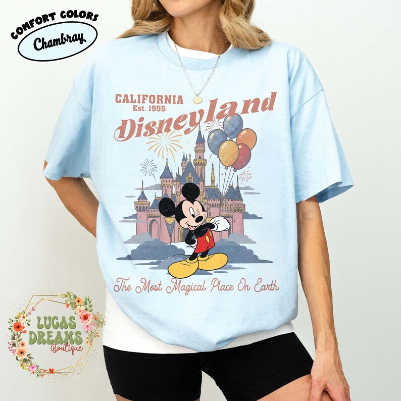May include: Light blue t-shirt featuring a graphic of Mickey Mouse in front of a castle with balloons. The text reads "CALIFORNIA Disneyland Est. 1955" and "The Most Magical Place On Earth". The shirt has the "Comfort Colors Chambray" logo.