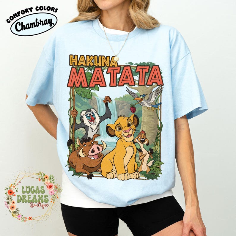 May include: Light blue t-shirt featuring the phrase "HAKUNA MATATA" in orange block letters, with a cartoon illustration of characters from The Lion King. The shirt has short sleeves and a crew neck. The brand name "Comfort Colors Chambray" is printed on the top left.