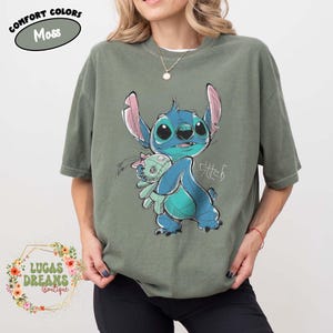 May include: A moss green short-sleeve t-shirt featuring a graphic illustration of the blue character Stitch holding a small doll. The design includes the text Stitch in a stylized script. A label in the corner reads Comfort Colors Moss.