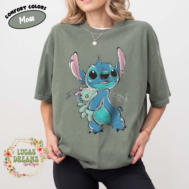 May include: A moss green short-sleeve t-shirt featuring a graphic illustration of the blue character Stitch holding a small doll. The design includes the text Stitch in a stylized script. A label in the corner reads Comfort Colors Moss.
