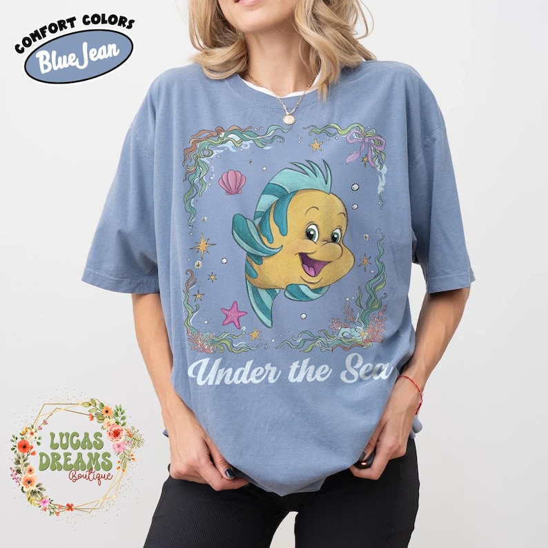 May include: Blue t-shirt with a graphic of a yellow and blue cartoon fish inside a decorative frame. The text "Under the Sea" is below the graphic. The shirt has the text "Comfort Colors Blue Jean" on the top left.