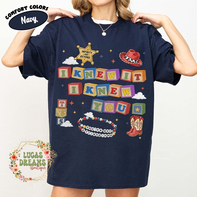May include: A navy blue short-sleeve t-shirt featuring a graphic print of colorful alphabet blocks spelling I KNEW IT I KNEW YOU. The design includes a gold sheriff star, a red cowboy hat, a beaded bracelet with the text YOU'VE GOT A FRIEND IN ME.