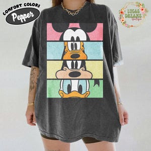 May include: Dark gray Comfort Colors t-shirt featuring a stacked graphic of Mickey Mouse, Goofy, and Donald Duck. The graphic is divided into horizontal color blocks of pink, blue, yellow, and green. The shirt has the text "Comfort Colors Pepper" on the upper left.