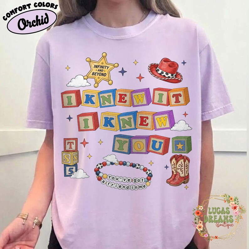 May include: A light purple t-shirt featuring a graphic print of colorful alphabet blocks spelling out I KNEW IT I KNEW YOU. The design includes a yellow sheriff star, a red cowboy hat, a pair of cowboy boots, and a beaded bracelet with text.