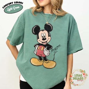 May include: A light green short-sleeve t-shirt featuring a distressed graphic of Mickey Mouse with the text Mickey Mouse. The shirt includes a label in the top left corner reading Comfort Colors Light Green and a floral logo in the bottom right.