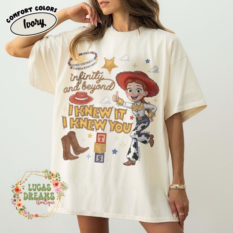 May include: An ivory t-shirt featuring a graphic print of the character Jessie from Toy Story. The design includes the text infinity and beyond, I KNEW IT, and I KNEW YOU, along with illustrations of a red cowboy hat, brown boots, and alphabet blocks.