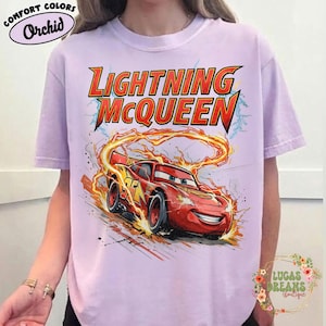 May include: A light purple t-shirt featuring a graphic of Lightning McQueen from the movie Cars. The design includes the text "Lightning McQueen" in red and yellow, with the race car surrounded by flames. The shirt has short sleeves.
