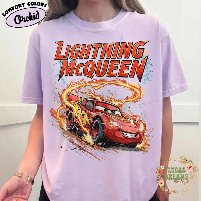 May include: A light purple t-shirt featuring a graphic of Lightning McQueen from the movie Cars. The design includes the text "Lightning McQueen" in red and yellow, with the race car surrounded by flames. The shirt has short sleeves.