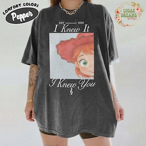 May include: A charcoal grey short-sleeve t-shirt featuring a graphic of a red-haired character wearing a red hat. The text reads EST 2026, I Knew It, and I Knew You with a treble clef symbol. The shirt has a crew neckline and a relaxed fit.