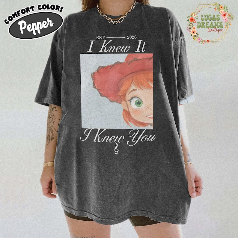 May include: A charcoal grey short-sleeve t-shirt featuring a graphic of a red-haired character wearing a red hat. The text reads EST 2026, I Knew It, and I Knew You with a treble clef symbol. The shirt has a crew neckline and a relaxed fit.