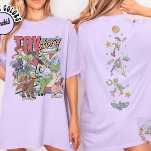 May include: A light purple t-shirt featuring a Toy Story graphic. The front of the shirt displays various characters with the text "Toy Story" and "Since 1995". The back shows Woody, Buzz Lightyear, and other characters.