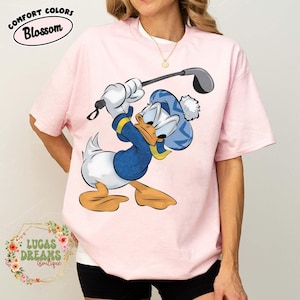 May include: Light pink t-shirt featuring a cartoon Donald Duck character in a blue golf outfit, swinging a golf club. The shirt has the text "COMFORT COLORS Blossom" at the top and "LUCAS DREAMS Boutique" at the bottom.