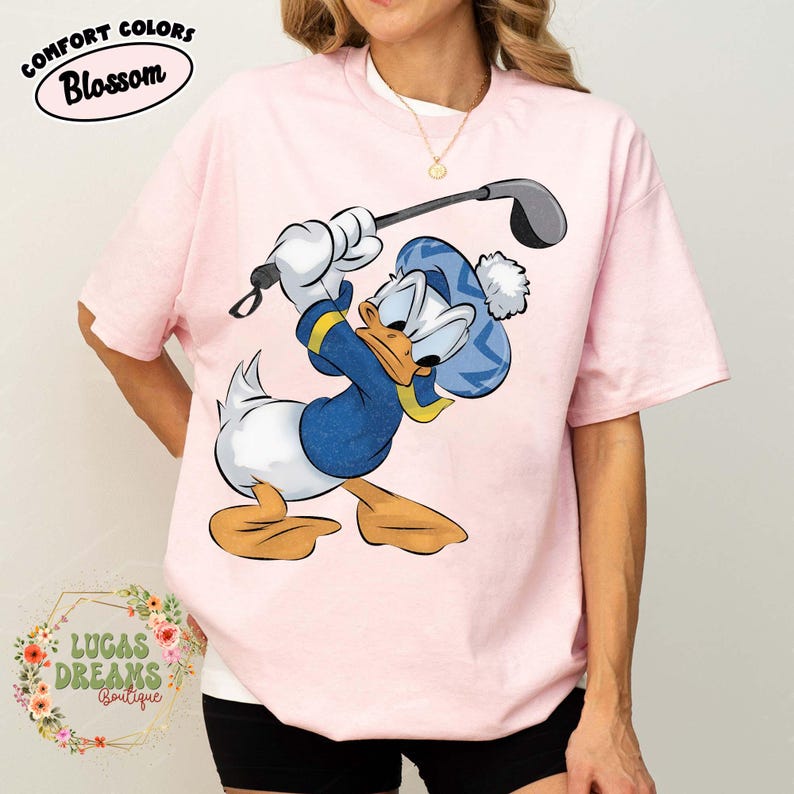 May include: Light pink t-shirt featuring a cartoon Donald Duck character in a blue golf outfit, swinging a golf club. The shirt has the text "COMFORT COLORS Blossom" at the top and "LUCAS DREAMS Boutique" at the bottom.
