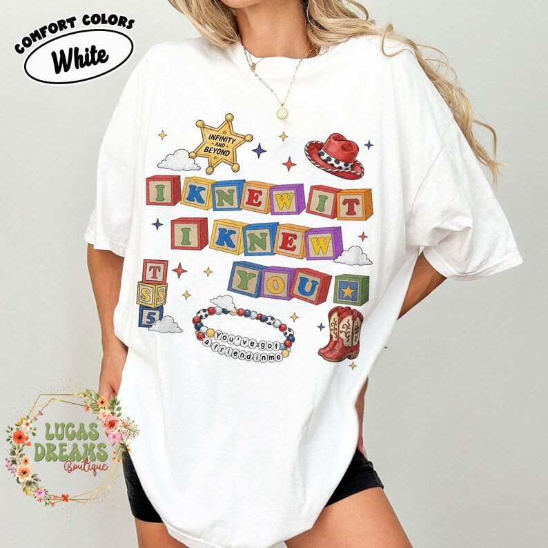 May include: A white t-shirt featuring a graphic print of colorful alphabet blocks spelling I KNEW IT I KNEW YOU, a sheriff star badge with the text INFINITY AND BEYOND, a cowboy hat, a pair of cowboy boots, and a beaded bracelet with the text YOU'VE GOT A FRIEND IN ME.