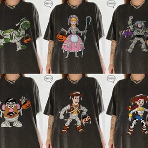May include: A collection of six dark gray t-shirts featuring various Toy Story characters dressed in mummy costumes and holding jack-o'-lanterns. The designs include Rex, Bo Peep, Buzz Lightyear, Mr. Potato Head, Woody, and Jessie.