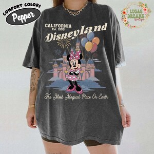 May include: Dark grey t-shirt featuring a Minnie Mouse graphic in front of a castle with balloons and fireworks. The shirt has the words "California Disneyland Est. 1955" and "The Most Magical Place On Earth". The shirt also has the words "Comfort Colors Pepper".