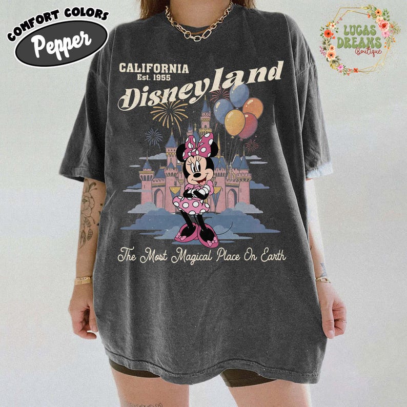 May include: Dark grey t-shirt featuring a Minnie Mouse graphic in front of a castle with balloons and fireworks. The shirt has the words "California Disneyland Est. 1955" and "The Most Magical Place On Earth". The shirt also has the words "Comfort Colors Pepper".